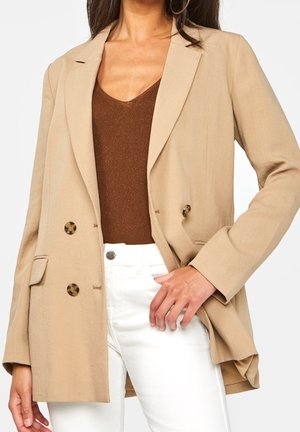 Vrouw met een beige double-breasted blazer, een bruin V-hals topje en witte broek, met één hand op haar heup.