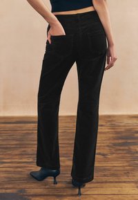 Pantaloni flared din corduroy negru, cu buzunare frontale și dorsale, având o suprafață texturată și o talie medie, asortați cu pantofi cu toc negri.