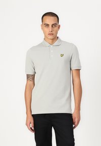 Světle šedé polo tričko s krátkým rukávem, dvouknoflíkovou légou a malým žlutým vyšívaným logem na levé části hrudi. Hladká textura.