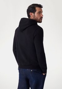 Sudadera negra con capucha, hecha de una mezcla de algodón, que presenta un corte relajado, puños y dobladillo acanalados, combinada con jeans de mezclilla azul.