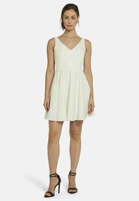 Weißes ärmelloses Kleid mit V-Ausschnitt, tailliertem Oberteil und schwingendem Rock. Trägt dazu schwarze, hochhackige Sandalen mit Riemchen. Glatte Textur, keine Muster.