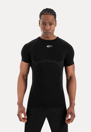 Hombre musculoso con el pelo trenzado, vistiendo una camiseta deportiva negra ajustada con un logo blanco, de pie frente a un fondo claro y sencillo.