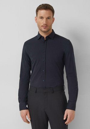 Mężczyzna ubrany w dopasowaną, ciemnogranatową koszulę na guziki i czarne spodnie, stojący na tle jednolitego jasnoszarego tła.