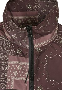 Veste marron à motifs avec des dessins de style bandana, un col montant et une fermeture éclair centrale avec un tire-zip noir.
