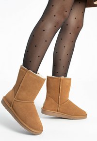 Tan suede boots with a shearling lining and a flat rubber sole. The design features a mid-calf height and stitching details along the seams.