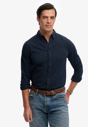 Mand med mørkt hår iført en navyblå knap-op-skjorte med opsmøgede ærmer, et brunt flettet bælte, blå jeans og et armbåndsur, stående foran en hvid baggrund.