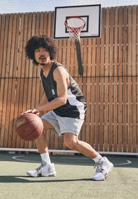 Black and white athletic tank top, gray shorts, and patterned sneakers. Holding a basketball, on a court with a wooden backdrop and hoop.