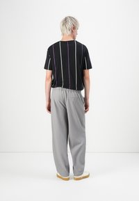 Person med kort blondt hår står med ryggen til, iført en sortstribet t-shirt, løse grå bukser og hvide sneakers med gummisåler.