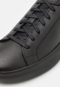 Sneaker in pelle nera con superficie testurizzata, dotato di lacci neri intrecciati, punta rotonda e suola in gomma con dettagli antiscivolo.