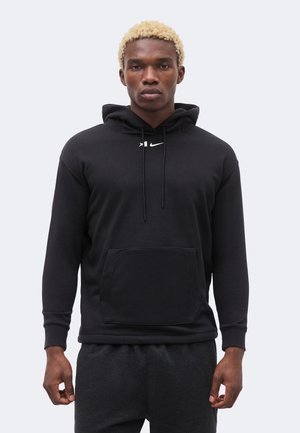 Άνδρας με κοντά ξανθά μαλλιά, φορώντας μαύρο φούτερ Nike με κορδόνια και μπροστινή τσέπη, στέκεται μπροστά σε απλό ανοιχτό φόντο.