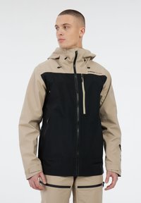 Man in een beige en zwarte hooded jack met ritszakken en beige broek met zwarte ritsdetails, kijkt naar links.