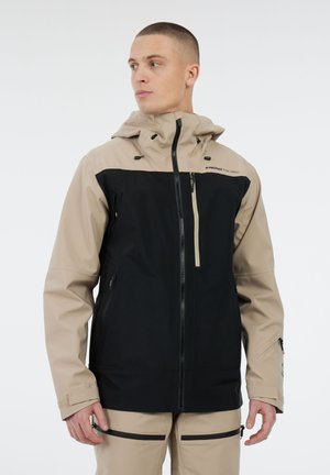 Man in een beige en zwarte hooded jack met ritszakken en beige broek met zwarte ritsdetails, kijkt naar links.