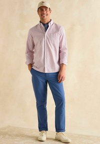 Pruhovaná červená a biela košeľa s gombíkmi, modré nohavice, béžová čiapka a biele tenisky. Pohodlný outfit s vyhrnutými rukávmi košele.