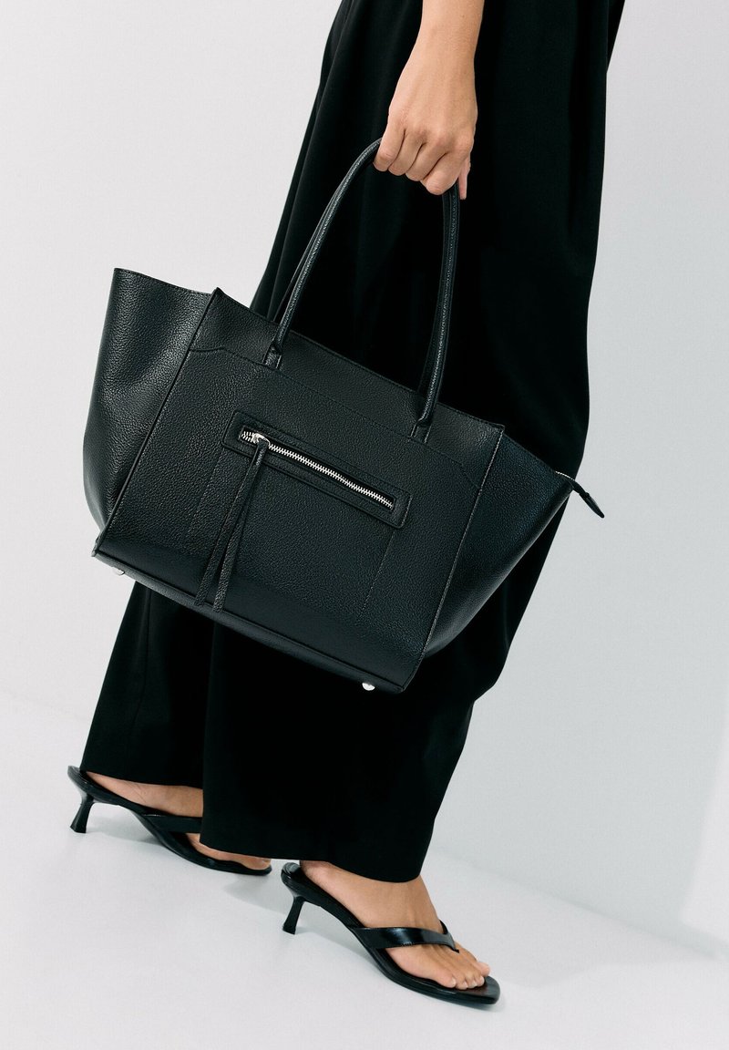 Personne portant un pantalon noir et des sandales à talons noires tenant un grand sac à main en cuir noir avec une poche zippée.