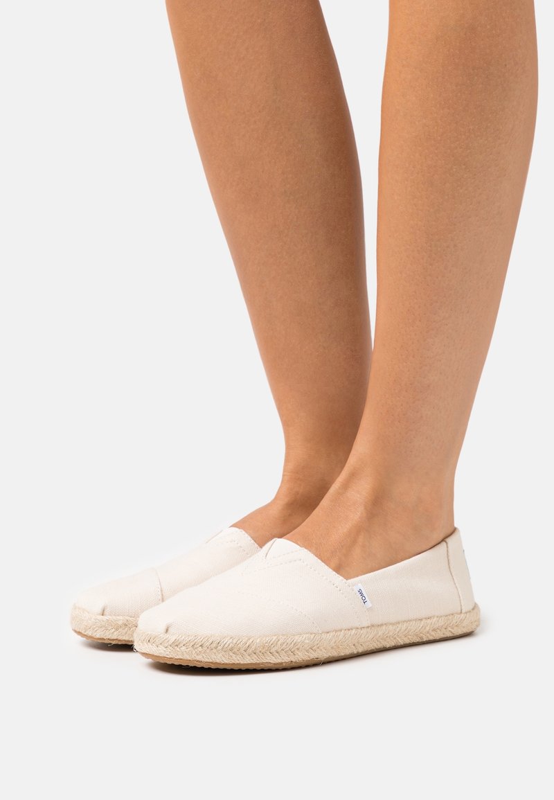Pieds portant des chaussures en toile blanches à enfiler avec des semelles en jute tressé sur un fond clair uni.