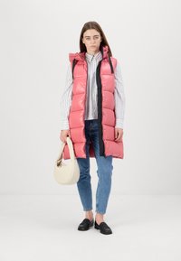 Gilet lungo imbottito rosa con cappuccio, dotato di zip frontale e dettagli neri. Il modello indossa una maglietta a righe, jeans blu e mocassini neri.