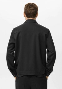 Moški stoji s hrbtom obrnjenim proti kameri, oblečen v črno jakno s kragnico in črne hlače, proti enobarvni beli ozadju.