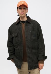 Joven con trenzas que lleva una gorra naranja, una chaqueta de cuadros marrón, un suéter marrón y pantalones cargo beige frente a un fondo simple.