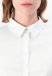 Camicia bianca con colletto, realizzata in tessuto morbido, caratterizzata da una chiusura frontale con bottoni trasparenti e un design pulito e minimale.