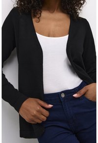 Femme portant un cardigan noir sur un haut blanc avec un jean bleu foncé, main ajustant le cardigan près de la taille.