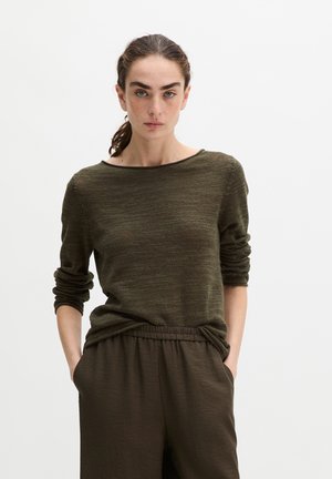 Frau mit braunen Haaren im Pferdeschwanz trägt einen langärmeligen olivgrünen Strickpullover und passende lockere Hose mit Händen in den Taschen.