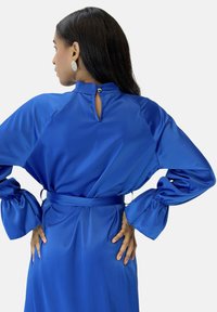 Abito blu in raso con maniche a sbuffo, collo alto e dettaglio posteriore a goccia con chiusura a bottone dorato, cintato in vita.