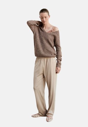 Woman wearing loose beige pants, brown off-shoulder sweater exposing pink lace strap, standing with one hand behind her neck.