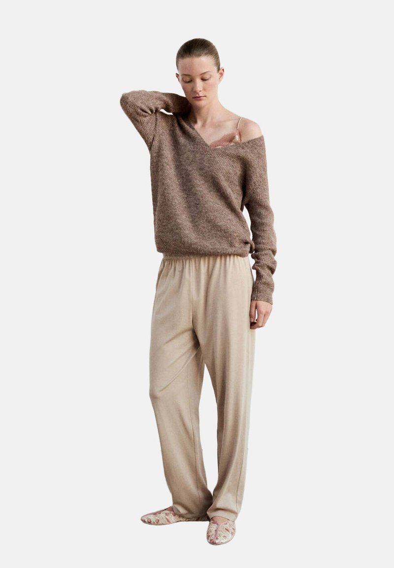 Femme portant un pantalon beige ample, un pull marron épaules dénudées laissant apparaître une bretelle rose en dentelle, debout avec une main derrière le cou.