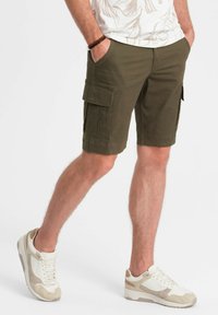 Shorts cargo vert olive avec deux poches latérales, en coton, conception légère et coupe droite, associés à des baskets beiges et blanches.