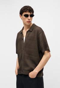 Νεαρός άνδρας με μαύρα γυαλιά ηλίου, καφέ υφασμένη κοντομάνικη πουκάμισο με κουμπιά και μαύρο παντελόνι, με τα χέρια στις τσέπες.