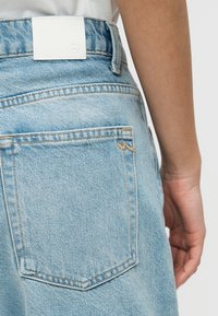 Τσέπη πίσω και μέση σε ανοιχτό μπλε τζιν με λευκή ετικέτα, φορεμένα από άτομο με ορατό χέρι και μπράτσο στη δεξιά πλευρά.