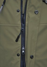 Grüne, wasserdichte Jacke mit einem zentralen schwarzen Reißverschluss, Druckknopfverschlüssen und einem strukturierten Stoff. Enthält einen Ziehling am Reißverschluss.