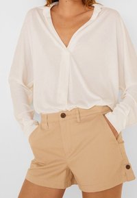 Blusa blanca de manga larga con escote en V y corte relajado, combinada con pantalones cortos beige claro que cuentan con detalle de botones y bolsillos laterales.