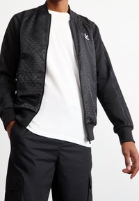 Svart bomberjacka med mönstrad framstycke, dragkedja och ribbade muddar, bärs över en vit t-shirt och svarta byxor.