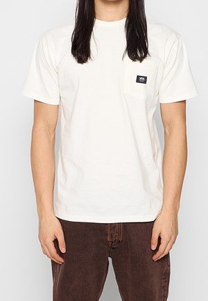 Hvid bomulds T-shirt med korte ærmer og en lomme foran, med et lille sort logo på lommen, parret med brune bukser.