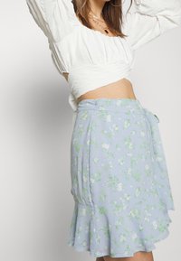 Světle modrá květinová zavinovací sukně s rozšířeným lemem, zdobená bílými a zelenými květinovými vzory. Vyrobena z jemné, lehké tkaniny.