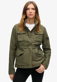 Oliven grøn jakke med lynlås og knaplukning foran, to brystlommer, indsnævret talje og stehals med kontrasterende detaljer.