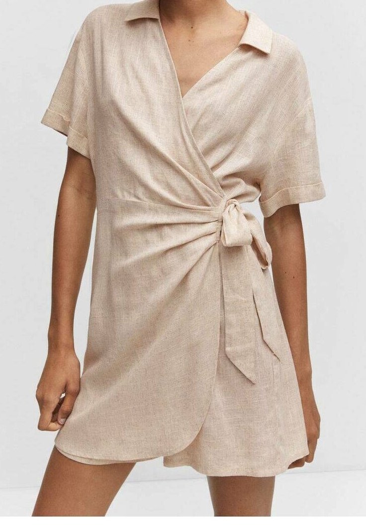 Robe portefeuille à manches courtes en lin beige clair. Elle présente un col, un détail noué à la taille et une fente latérale pour faciliter les mouvements.