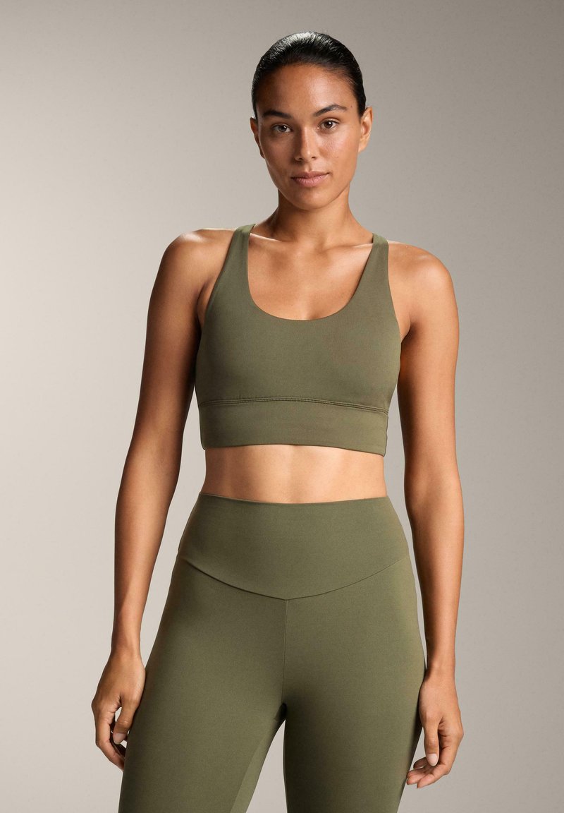 Športni top v olivno zeleni barvi z izrezom v obliki sklede in širokimi naramnicami, kombiniran z visokim pasom ujemajočih pajkic. Gladek, raztegljiv material z minimalnimi šivi.