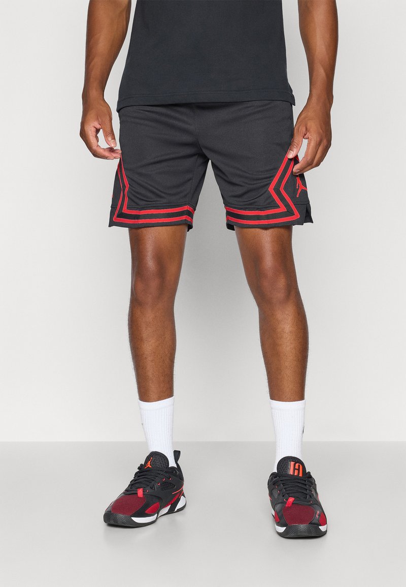 Mann som har på seg svarte treningsshorts med røde striper og svarte og røde joggesko, står mot en ensfarget bakgrunn.