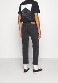 Schwarzes Grafik-T-Shirt mit Bergdesign und weißen Akzenten, kombiniert mit dunkelgrauen Jeans, weißen Socken und schwarzen Schuhen.