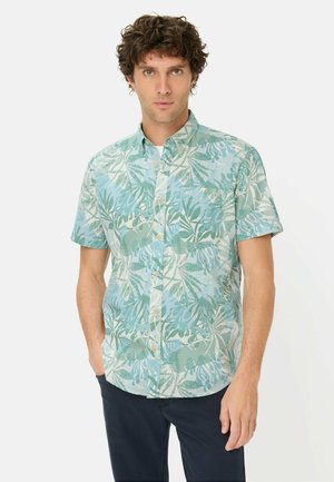 Mężczyzna w koszuli na guziki z krótkim rękawem z zielonym i niebieskim liściastym wzorem, stojący z jedną ręką w kieszeni na jednolitym tle.