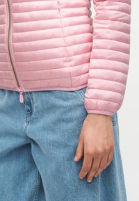 Κοντινό πλάνο ενός ατόμου που φοράει ροζ μπουφάν με καπιτονέ ύφασμα και ανοιχτό μπλε τζιν παντελόνι, με το δεξί του χέρι χαλαρό δίπλα στο σώμα.