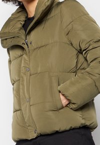 Olivgrön vadderad jacka med hög krage, dragkedja och tryckknappar. Har quiltad textur och sidofickor.