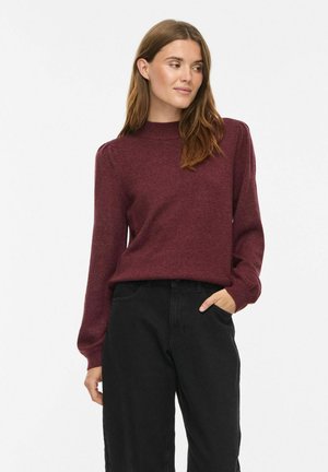 Donna che indossa un maglione bordeaux a maniche lunghe e pantaloni neri, in piedi con una mano in tasca, guardando di lato.