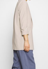 Blazer beige avec manches trois-quarts et coupe décontractée, doté d'une poche avant unique. Associé à un pantalon bleu ample.