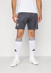 Un atlet masculin îmbrăcat cu pantaloni scurți gri de fotbal Arsenal, șosete albe Adidas până la genunchi cu dungi negre și ghete albe, stând pe un fundal simplu.