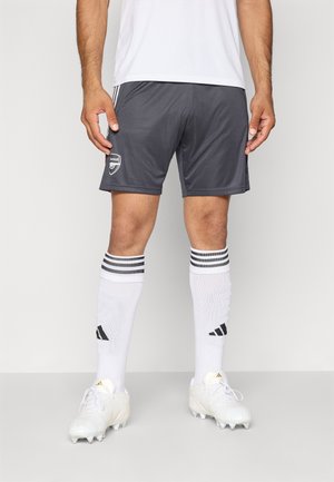 Mandlig atlet iført grå Arsenal fodboldshorts, hvide Adidas knæhøje sokker med sorte striber og hvide fodboldstøvler, stående på ensfarvet baggrund.