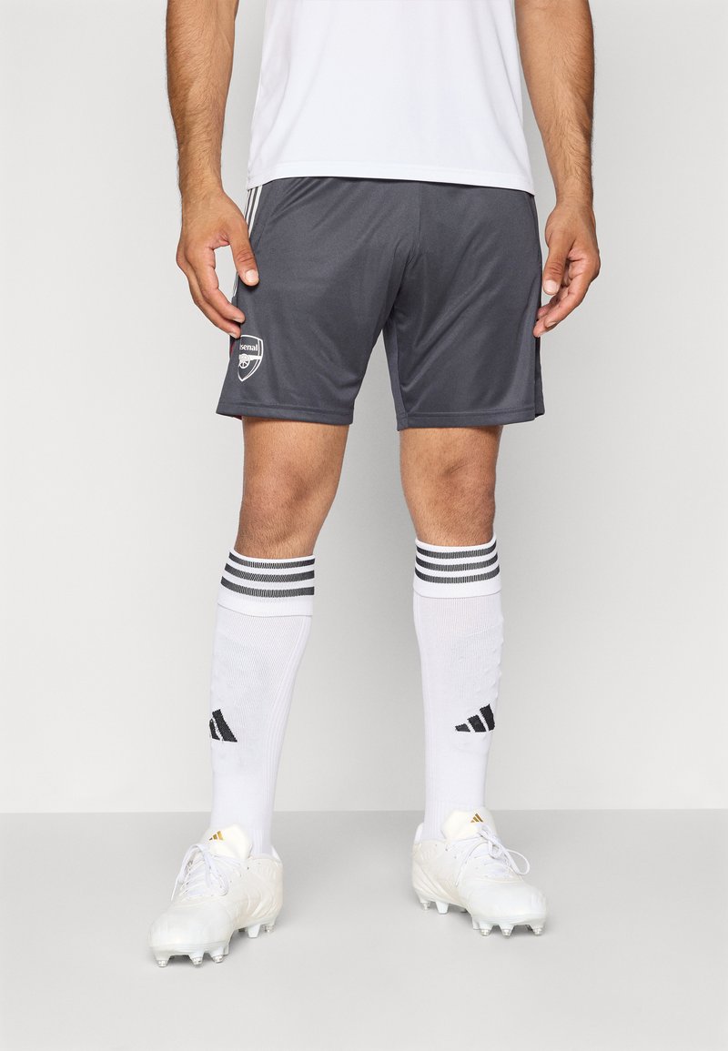 Un atlet masculin îmbrăcat cu pantaloni scurți gri de fotbal Arsenal, șosete albe Adidas până la genunchi cu dungi negre și ghete albe, stând pe un fundal simplu.