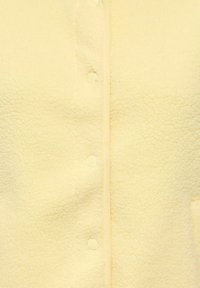 Rumena flis oblačilo z teksturirano površino, s zapiranjem na gumbe in kontrastnim obrobljenjem ob robovih.